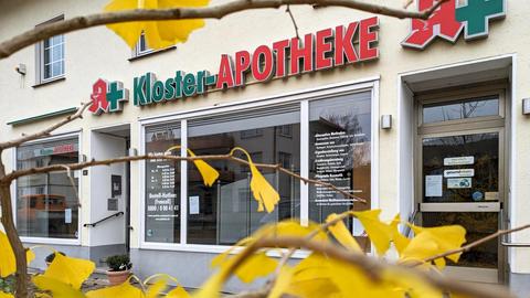 Außenansicht einer Apotheke. Schaufenster und Schriftzug "Kloster-Apotheke" mit Apothekenzeichen und Kreuz daneben. 