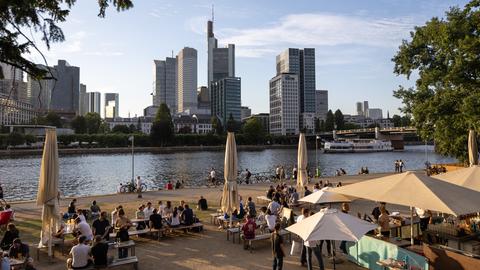 Das Bild zeigt ein Café am Main. Menschen sitzen auf Terrassenmöbeln auf einem Platz, im Hintergrund ist die Skyline zu sehen.