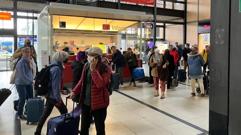 Reisende stehen in einer Bahnhofshalle.