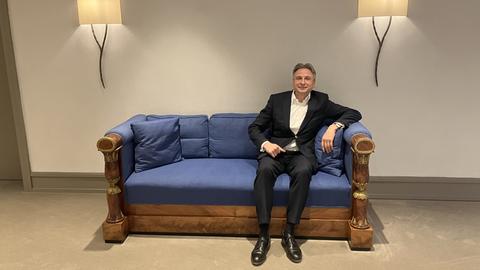 Ein Mann im Anzug sitzt auf einem alten blauen Sofa. Es ist der Vorstandssprecher des Bankhauses Metzler