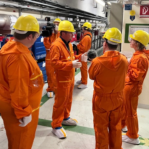 Eine Gruppe von Menschen in orangfarbenen Schutzanzügen und Helmen steht in einem Raum mit vielen Rohren und Technik.