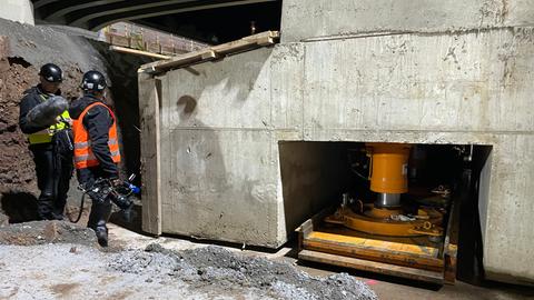 Zwei Bauarbeiter mit Helm stehen an einem Betonfuß der Brücke. Zu sehen sind orange Hydraulikstempel, die das Brückenbauwerk anheben.