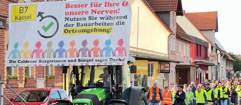 Ein Traktor mit einem Schild mit der Aufschrift "Besser für Ihre und unsere Nerven! Nutzen Sie während der Bauarbeiten die Ortsumgehung!", dahinter Menschen in Warnwesten