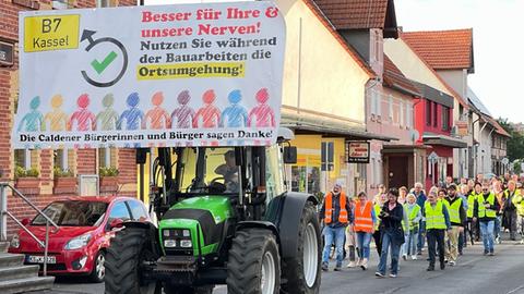 Ein Traktor mit einem Schild mit der Aufschrift "Besser für Ihre und unsere Nerven! Nutzen Sie während der Bauarbeiten die Ortsumgehung!", dahinter Menschen in Warnwesten