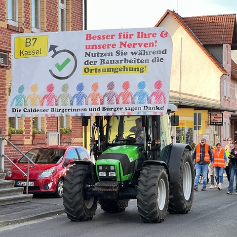 Ein Traktor mit einem Schild mit der Aufschrift "Besser für Ihre und unsere Nerven! Nutzen Sie während der Bauarbeiten die Ortsumgehung!", dahinter Menschen in Warnwesten