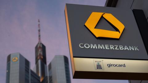 Im Bildvordergrund ein Commerzbank-Schild an einer Filiale in Frankfurt, im Hintergrund die Hochhäuser der Commerzbank.