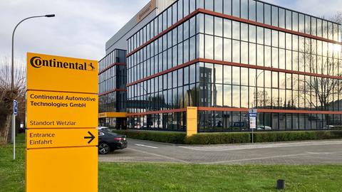 Bürogebäude mit verspiegelter Glasfassade, in welcher sich gelbliches Licht reflektiert, in der Außenansicht. Im Bildvordergrund ein orangfarbenes Schild mit der Aufschrift "Continental-Standort Wetzlar-Eingang"