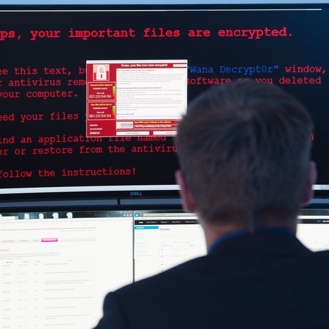 Ein Mann sitzt vor mehreren Computerbildschirmen. Auf einem steht der englische Text: "Ooops, your important files have been encrypted" zu lesen. 