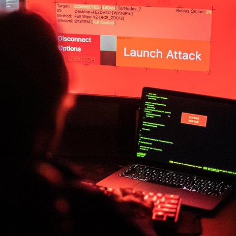 Eine Person, im Dunkeln von hinten fotografiert, sitzt vor leuchtenden Bildschirmen. Auf einem ist der Schriftzug "Launch Attack" zu lesen.