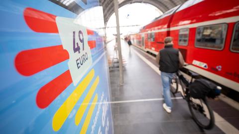 Foto eines Bahnsteigs: links eine Anzeigetafel, auf der "49 Euro" steht, rechts eine rote Regionalbahn, in der Mitte auf dem Bahnsteig eine Person mit einem Rad.
