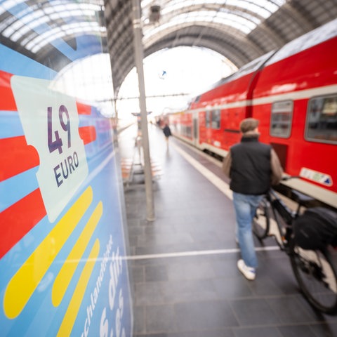 Foto eines Bahnsteigs: links eine Anzeigetafel, auf der "49 Euro" steht, rechts eine rote Regionalbahn, in der Mitte auf dem Bahnsteig eine Person mit einem Rad.