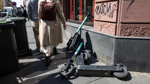 Zwei E-Scooter liegen auf einem Bürgersteig in der Nähe von Mülltonnen und blockieren den Weg