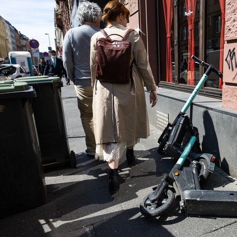 Zwei E-Scooter liegen auf einem Bürgersteig in der Nähe von Mülltonnen und blockieren den Weg