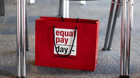 Eine Papiertüte mit der Aufschrift "Equal Pay Day" steht unter einem Bürostuhl