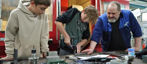 Zwei Schüler schrauben Metallteile, ein Mitarbeiter steht daneben.