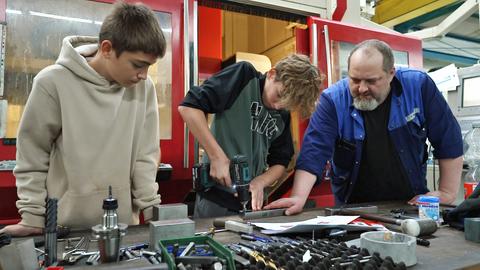 Zwei Schüler schrauben Metallteile, ein Mitarbeiter steht daneben.