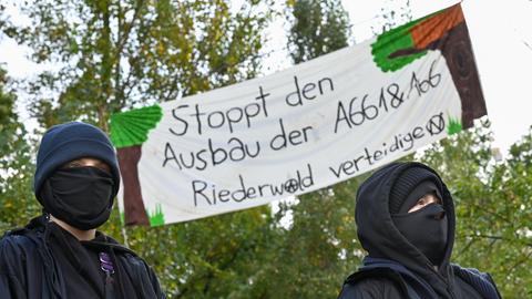 Zwei Aktivistinnen, hinter denen ein Plakat mit der Aufschrift "Stoppt den Ausbau der A661 & A66. Riederwald verteidigen" an Bäumen hängt.