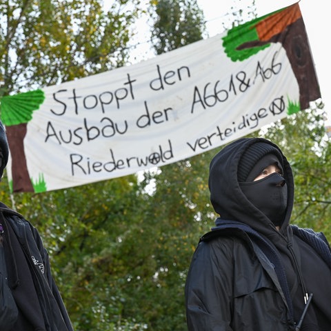 Zwei Aktivistinnen, hinter denen ein Plakat mit der Aufschrift "Stoppt den Ausbau der A661 & A66. Riederwald verteidigen" an Bäumen hängt.