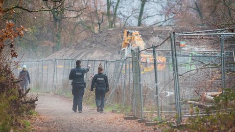 Zwei Polizeibeamte gehen an einem Bauzaun entlang, hinter welchem ein Bagger, viel geräumte Erde und einige Bäume zu sehen sind.