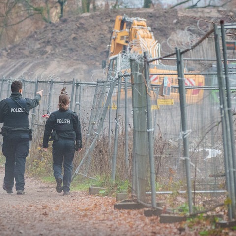 Zwei Polizeibeamte gehen an einem Bauzaun entlang, hinter welchem ein Bagger, viel geräumte Erde und einige Bäume zu sehen sind.