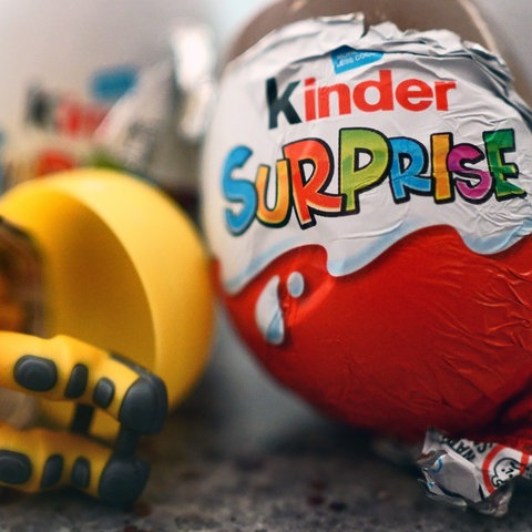 Ein kleines Spielzeug in einer gelben Kapsel liegt neben einem Ferrero-Überraschungsei.