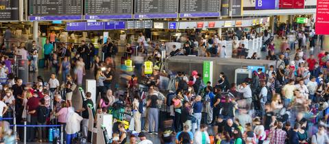Viele Menschen mit Koffern stehen in einer großen Halle, dahiner eine Anzeigetafel für Flüge