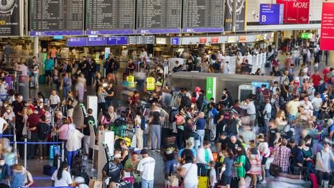 Viele Menschen mit Koffern stehen in einer großen Halle, dahiner eine Anzeigetafel für Flüge