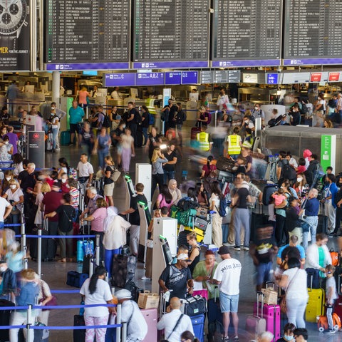 Viele Menschen mit Koffern stehen in einer großen Halle, dahiner eine Anzeigetafel für Flüge