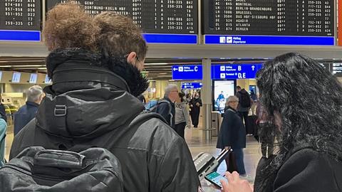 Zwei Frauen stehen vor einer Anzeigetafel am Flughafen Frankfurt, beide sind von hinten fotografiert.