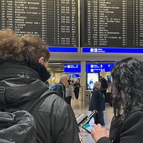 Zwei Frauen stehen vor einer Anzeigetafel am Flughafen Frankfurt, beide sind von hinten fotografiert.