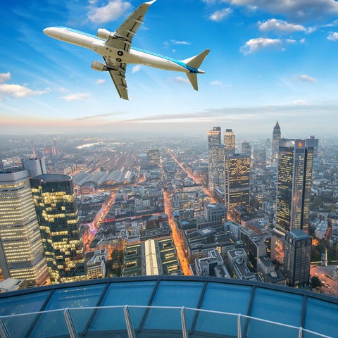 Ein Flugzeug am leuchtend blauen Himmel, darunter ragen die beleuchteten Hochhäuser der Skyline auf.