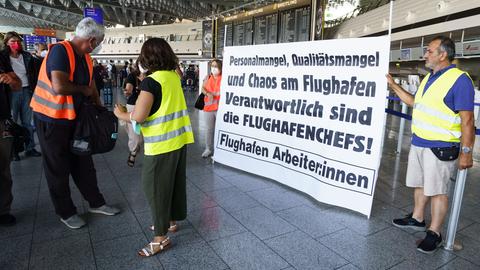 Mehrere Menschen mit Warnwesten stehen in der Abflughalle des Flughafens, zwei von ihnen halten ein Transparent mit der Aufschrift:"Personalmangel, Qualitätsmangel und Chaos am Fluhafen -Verantwortlich sind die Flughafenchefs!"