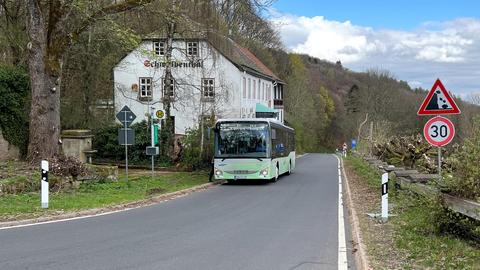 Ein Bus steht an einer Haltestelle auf einer Landstraße. Neben der Haltstelle ein weißes Gasthaus. Drumherum Landschaft.