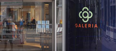 Gläserene Eingangstüren einer Galeria-Filiale mit einem Galeria-Logo auf einer Säule daneben. 