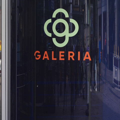 Gläserene Eingangstüren einer Galeria-Filiale mit einem Galeria-Logo auf einer Säule daneben. 