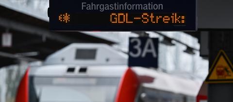 Das Bild zeigt eine Anzeigetafel an einem Bahngleis, auf der steht: GDL-Streik. Im Hintergrund sind eine S-Bahn zu sehen und eine Frau in hellblauer Winterjacke zu sehen.