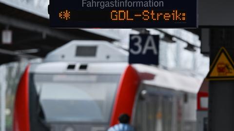 Das Bild zeigt eine Anzeigetafel an einem Bahngleis, auf der steht: GDL-Streik. Im Hintergrund sind eine S-Bahn zu sehen und eine Frau in hellblauer Winterjacke zu sehen.