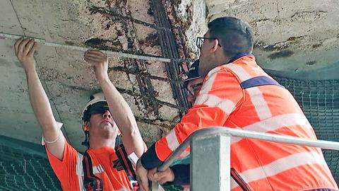 Drei Männer in neonfarbener Arbeitskleidung stehen unter einer Betondecke und begutachten eine marode Stelle, an welcher Beton abgebröckelt ist und bewehrende Stahlstangen offen liegen.