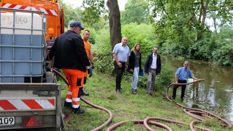 Männer und Frauen stehen neben einem Tank, aus dem ein Schlauch in einen Graben führt.