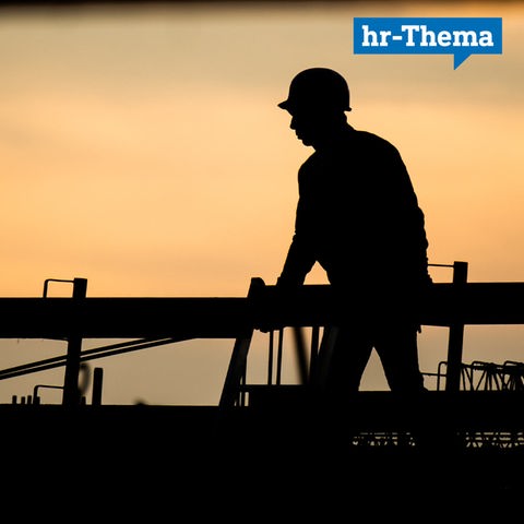 Ein Bauarbeiter in der Dämmerung gegen das Licht fotografiert, so dass fast nur eine schwarzer Scherenschnitt eines Mannes mit Helm vor einem orangfarbenen Hintergrund zu sehen ist.