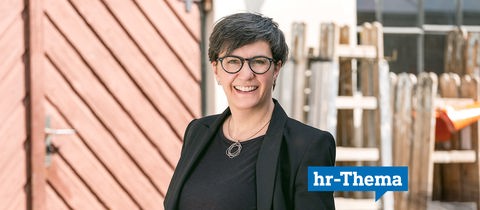 Die Präsidentin der Handwerkskammer steht vor dem Gebäude eines Handwerkbetriebs und lächelt in die Kamera. Auf dem Foto klebt ein Label mit der Aufschrift "hr Thema".