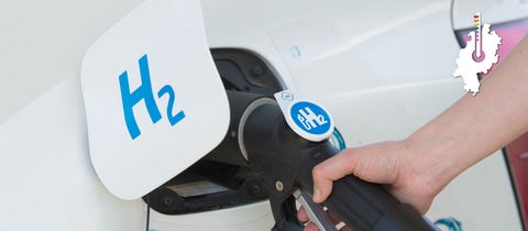 Eine Hand hält eine Tankpistole, welche in eine Zapfstelle mit der Bezeichnung "H2" gesteckt wurde.