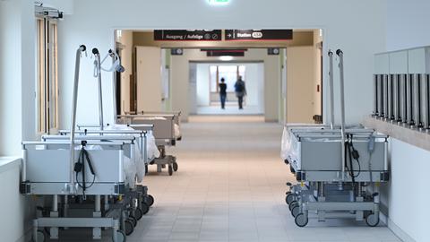 Ein Flur im Klinikum, links und rechts stehen leere Patientenbetten mit Plastikabdeckung. Im Hintergrund gehen zwei Menschen (unscharf).