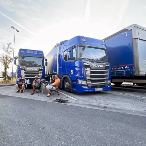 Vier Männer sitzen auf Campingstühlen vor Lastwagen auf einer Raststätte.