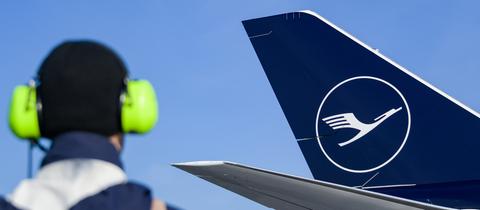 Foto: Im Vordergrund unscharf ein Mensch mit gelbem Lärmschutz auf den Ohren. Im Hintergund scharf Teil eines Flugzeuges mit dem Lufthansa-Logo.