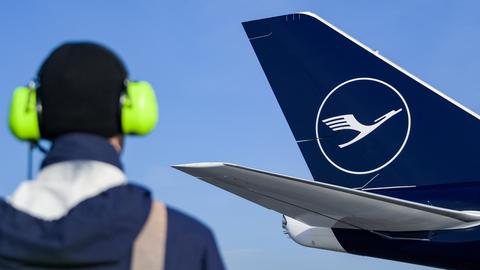 Foto: Im Vordergrund unscharf ein Mensch mit gelbem Lärmschutz auf den Ohren. Im Hintergund scharf Teil eines Flugzeuges mit dem Lufthansa-Logo.