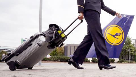 Aus der Froschperspektivr fotografiert: Unterkörper eines gehenden Menschen in dunkelblauer Uniform von der Seite, der einen Rollkoffer hinter sich herzieht. Im Bildhintergrund Teil eines Flugzeuges mit Lufthansa-Logo.