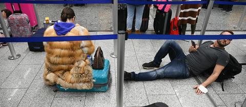 Eine Frau sitzt auf ihrem Koffer, ein Mann liegt auf dem Boden zwischen blauen Absperrbändern.