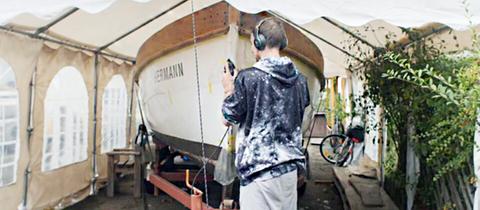 Ein junger Mann in Kapuzenpulli und mit Kopfhörer steht vor einem Boot und arbeitet daran.