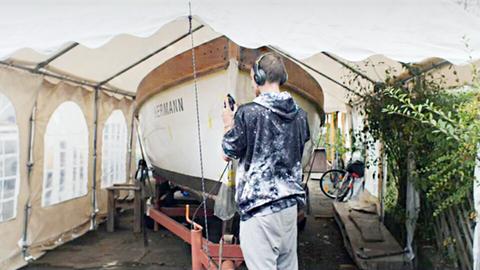 Ein junger Mann in Kapuzenpulli und mit Kopfhörer steht vor einem Boot und arbeitet daran.